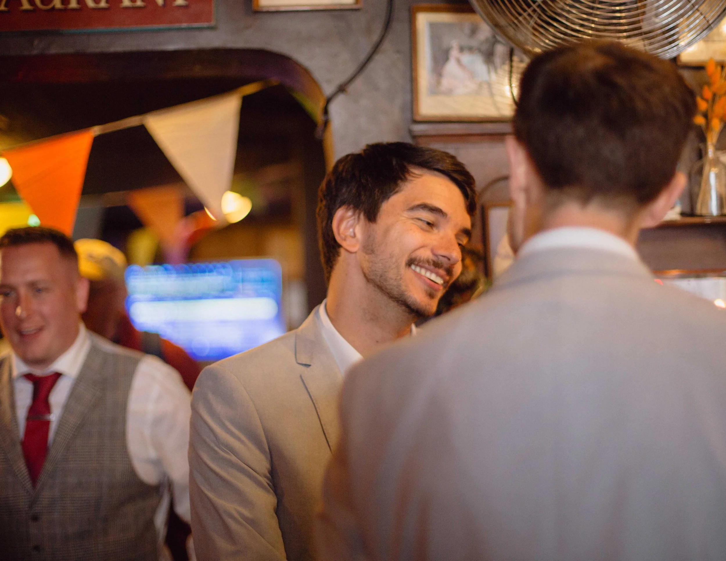 Three men in suits are at a social gathering in a dimly lit room with festive decorations, including bunting and warm lighting. One man is smiling and engaged in conversation, while another is blurred in the background.