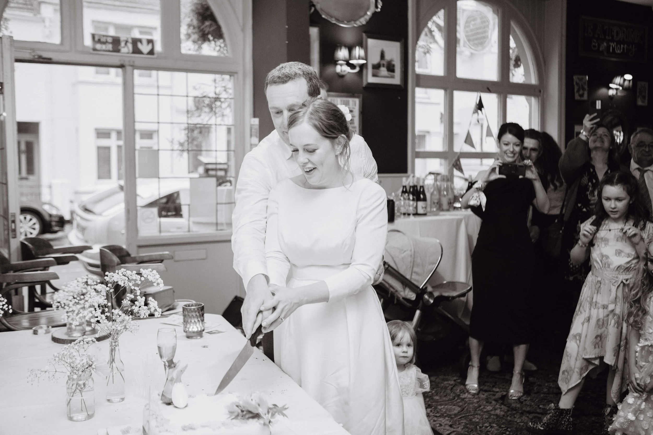 A bride and groom cutting a wedding cake in a celebration with guests taking photos and watching.
