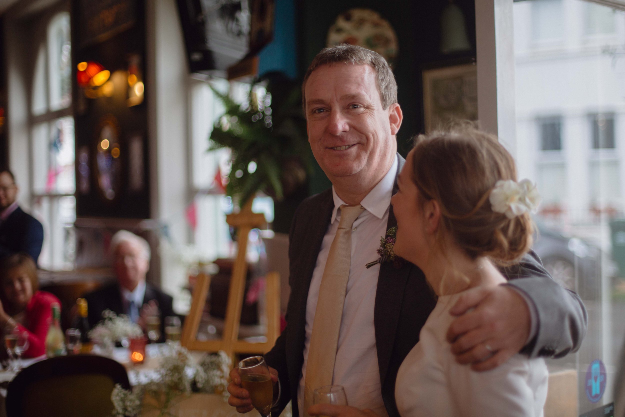 A man in a suit holding a glass of champagne, smiling, with a woman with a flower in her hair, at a social gathering in a decorated venue with large windows and other guests seated.