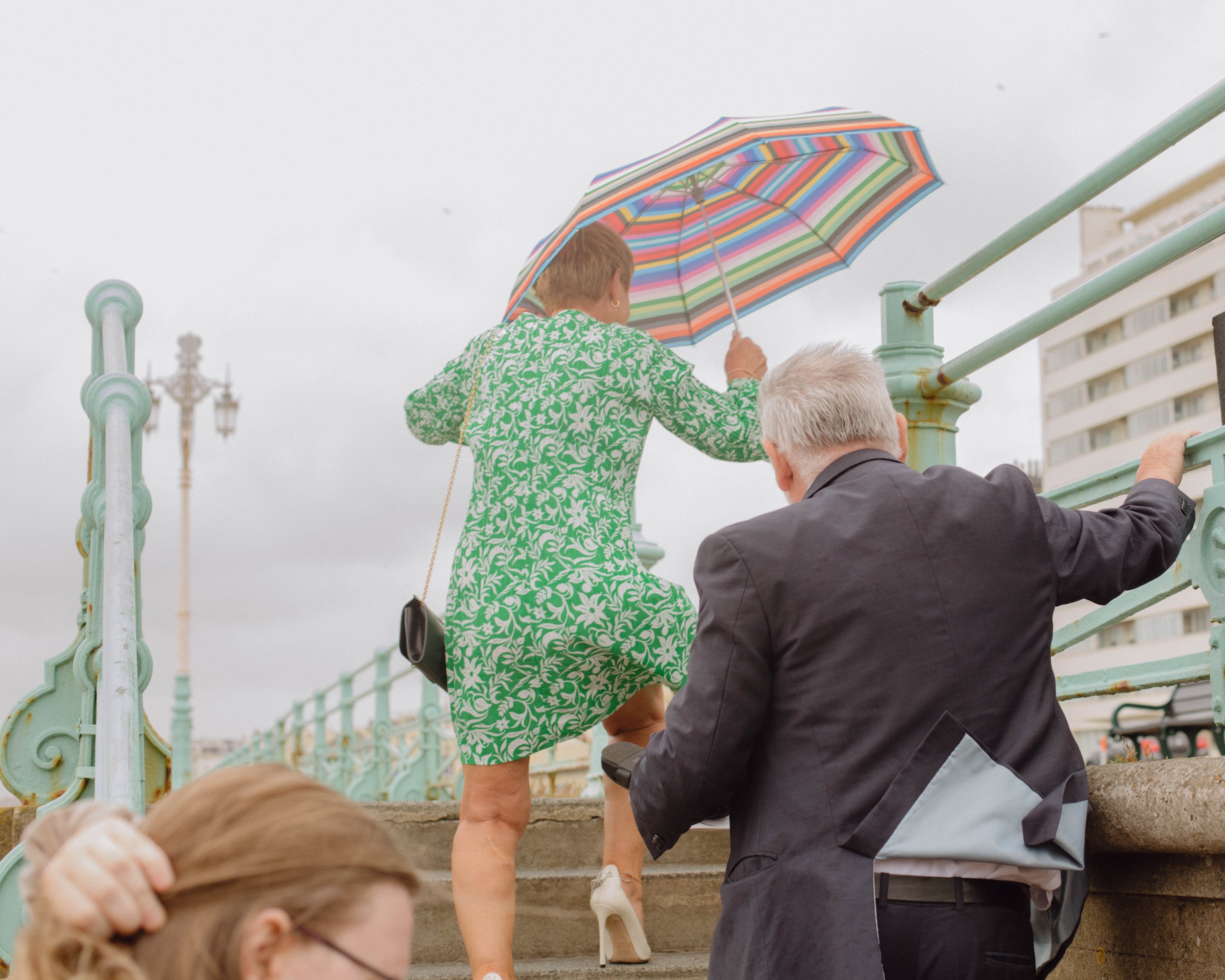 A woman in a green patterned dress carrying a rainbow-striped umbrella, walking up stone steps while a man in a dark suit assists her, during a cloudy day near a railing.