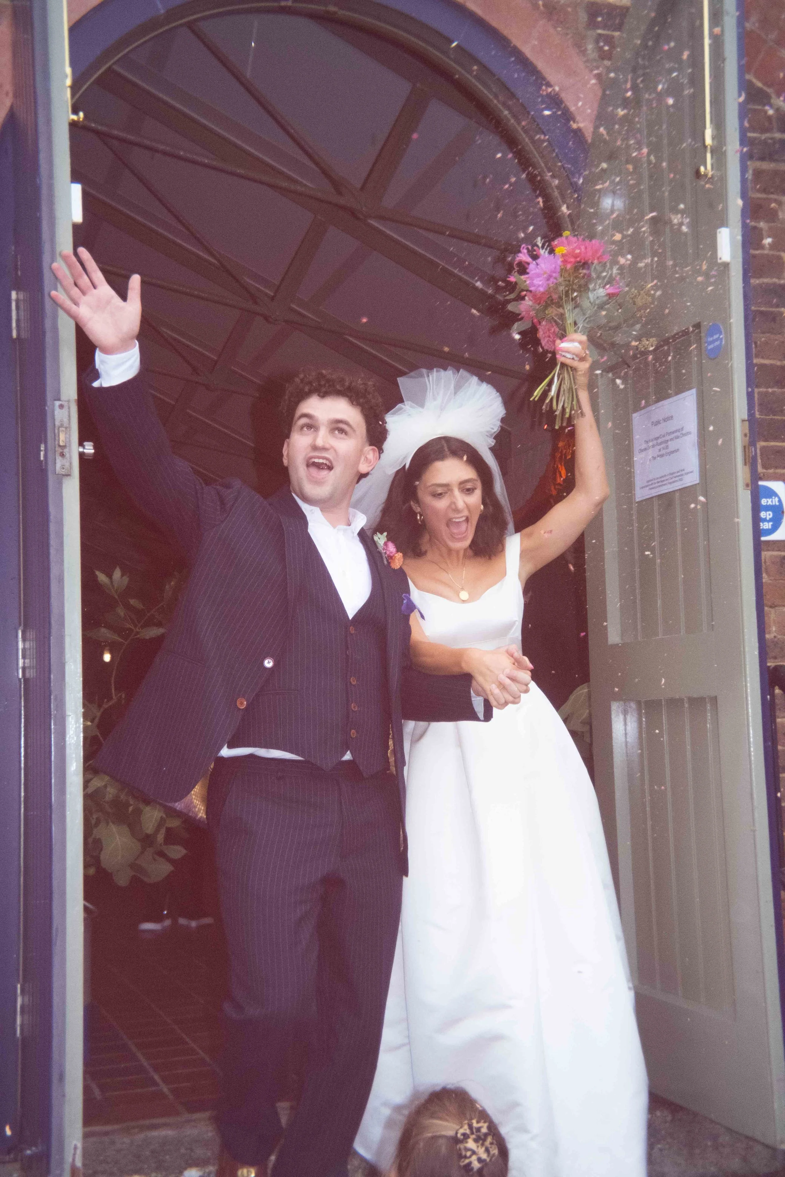 A newlywed couple celebrating as they exit a building, with the bride holding a bouquet of flowers and the groom smiling and waving.