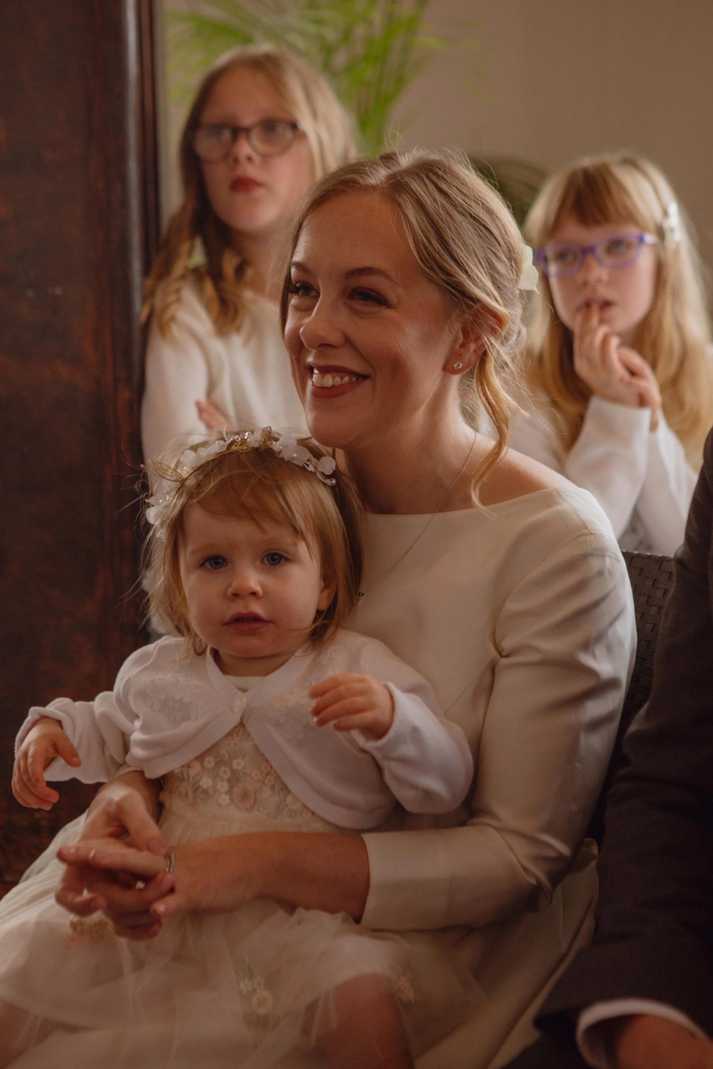 A woman holding a young girl, with two girls standing in the background, all dressed in white. They are inside a room with a wooden panel and a plant in the background.