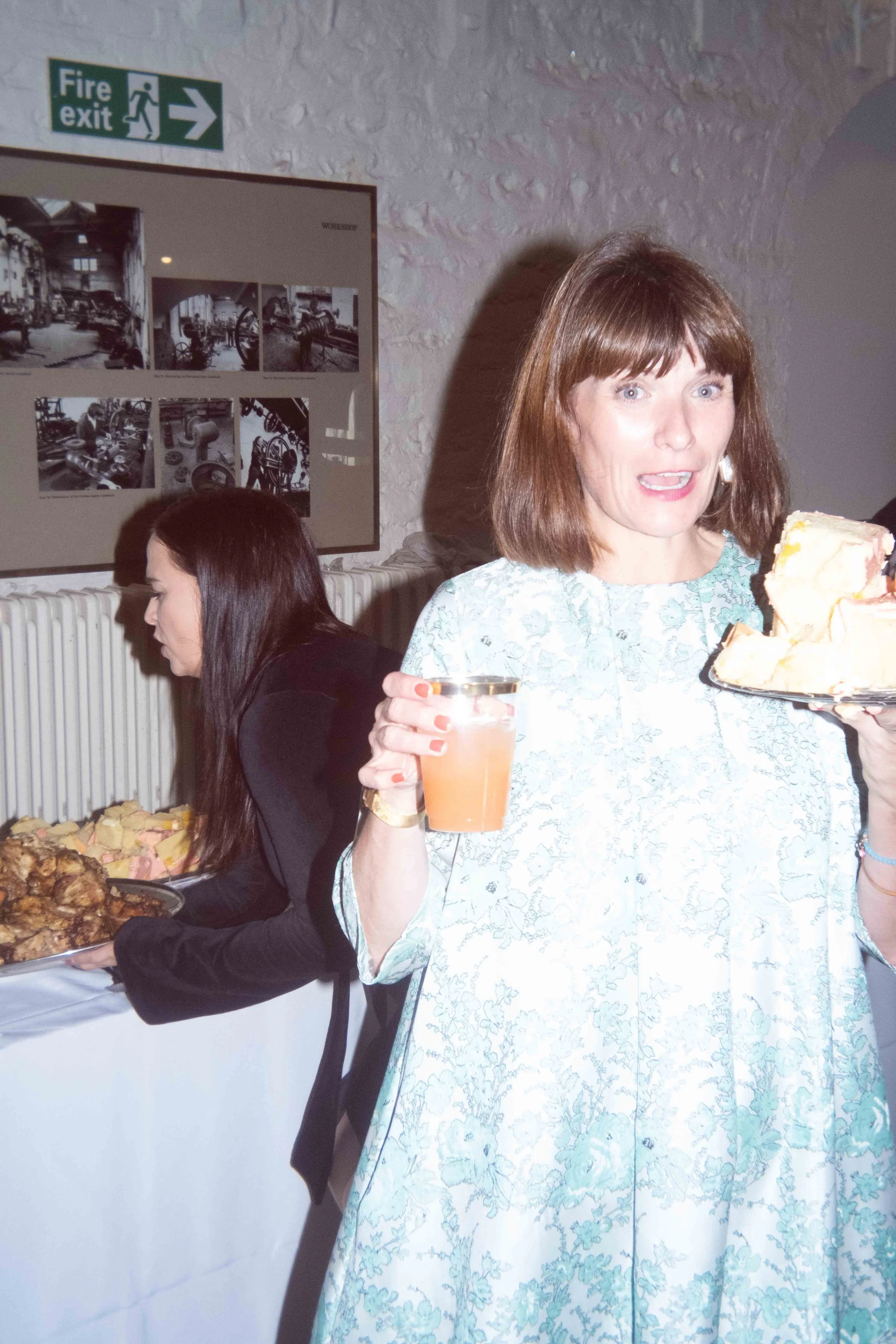 Woman holding a glass of beverage and a plate with a large slice of cake at a social gathering.