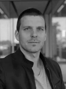 Black and white portrait of a Tom Fokkens-Ancery  with short hair, wearing a dark jacket over a light shirt, standing indoors near large windows.