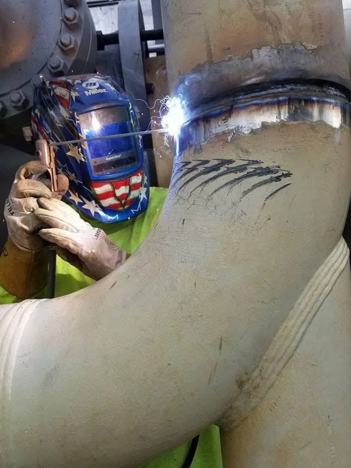 A worker wearing a welding helmet with an American flag design is welding a large pipe.