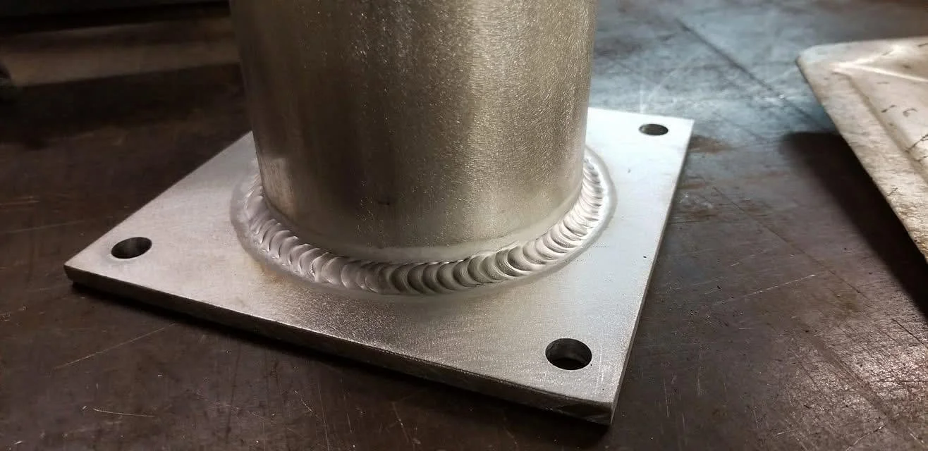 Close-up of a metallic pipe welded onto a square metal plate with four holes at the corners, resting on a wooden work surface.