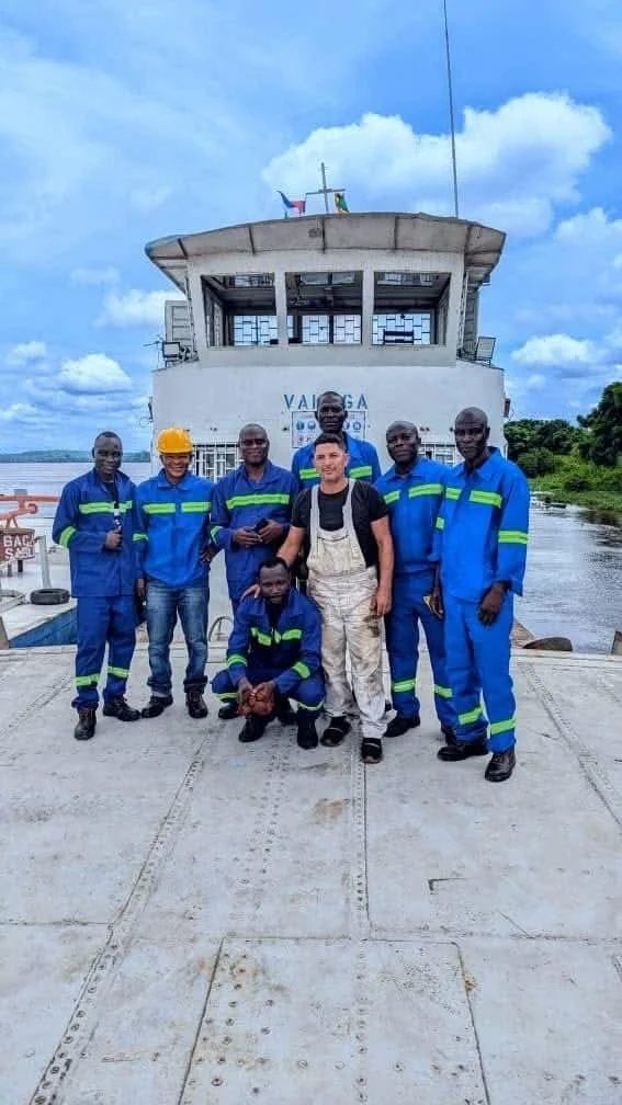 Group of seven men, some in blue work uniforms with fluorescent stripes, standing on a dock in front of a boat. One man is in white overalls, and one is wearing a yellow safety helmet. The boat has the name '_VALGRA_' on it and is on a river with greenery in the background under a partly cloudy sky.