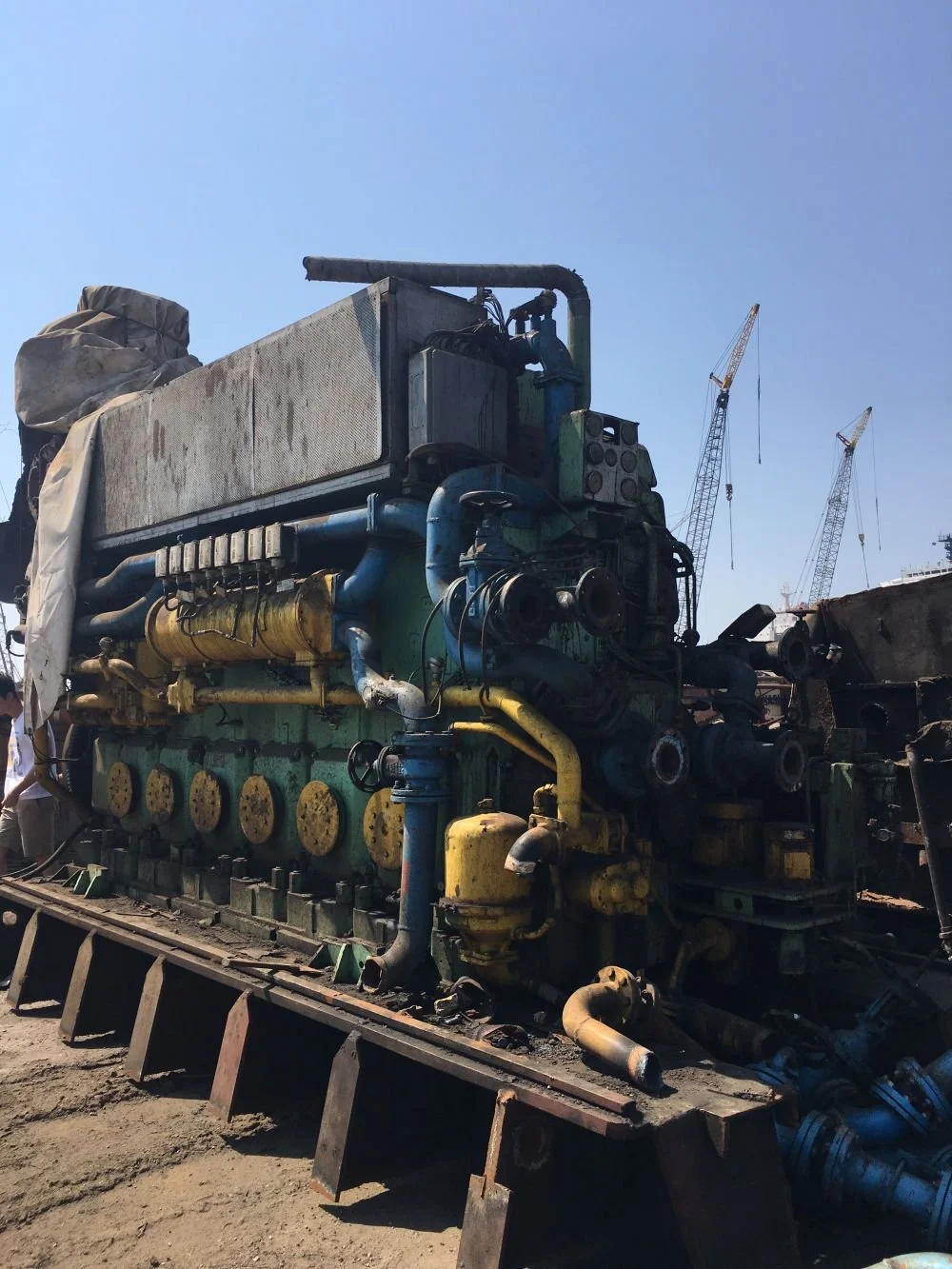 An old, large diesel engine with various pipes and components, sitting on a dirt surface at a construction or industrial site with cranes in the background.