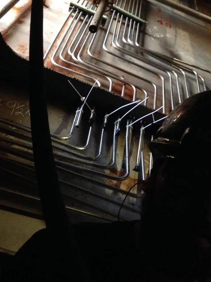A collection of assorted L-shaped metal hooks arranged on a dark surface, with some wires attached to a few of the hooks, and a portion of a black helmet and dark clothing visible on the right side.