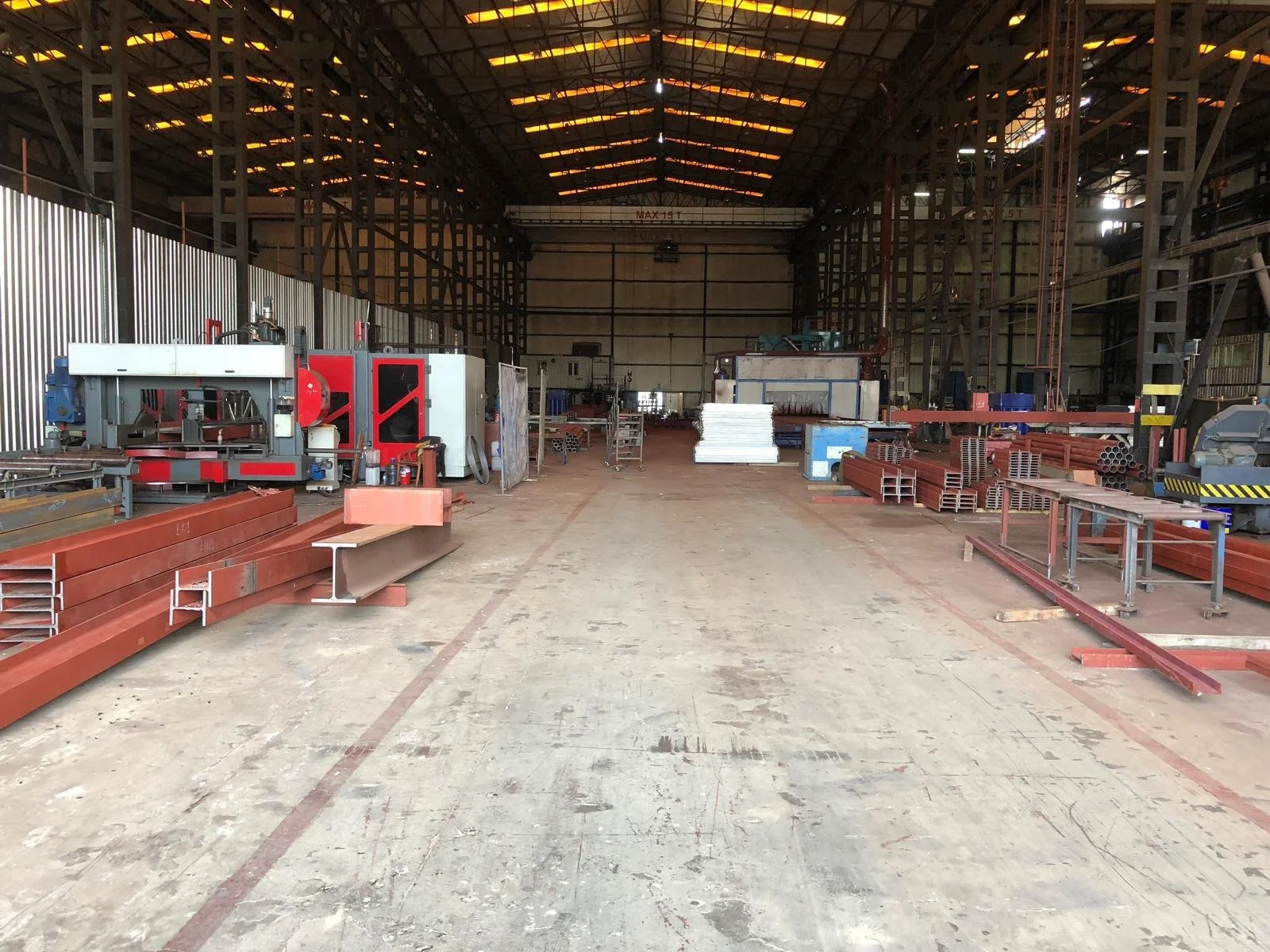 An empty industrial warehouse with various pieces of machinery, metal pipes, and construction materials scattered throughout. The warehouse has a high ceiling with exposed beams and some gaps letting in light.