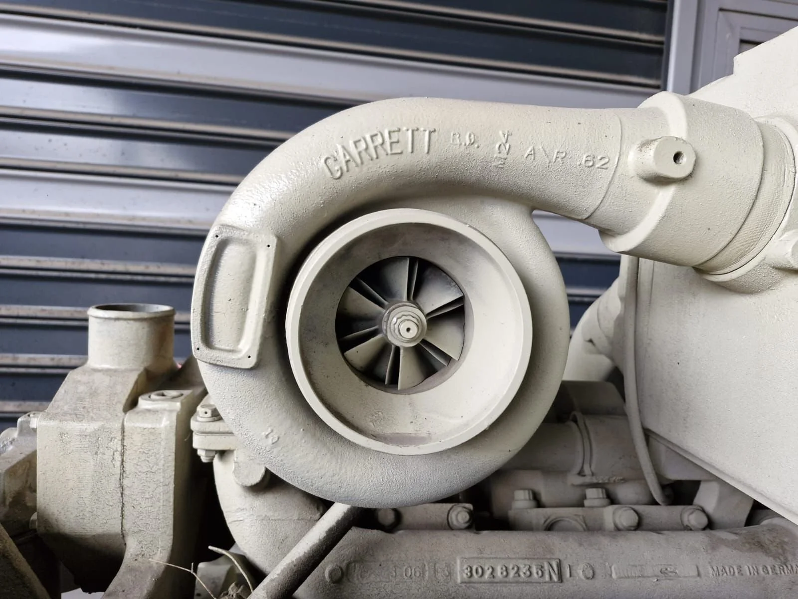 Close-up of a turbocharger with a Garrett label, showing the turbine wheel inside the housing, mounted on an engine part against a gray metal background.