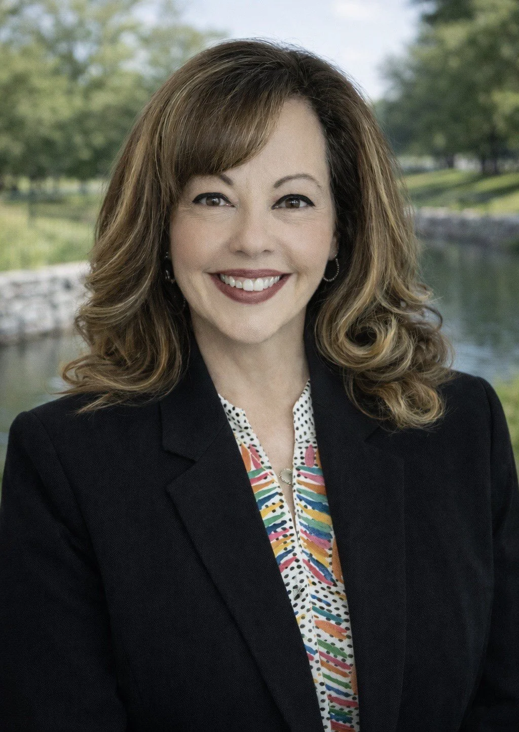 A woman with shoulder-length wavy hair, smiling, wearing a gray blazer and light blue shirt, standing indoors near windows.