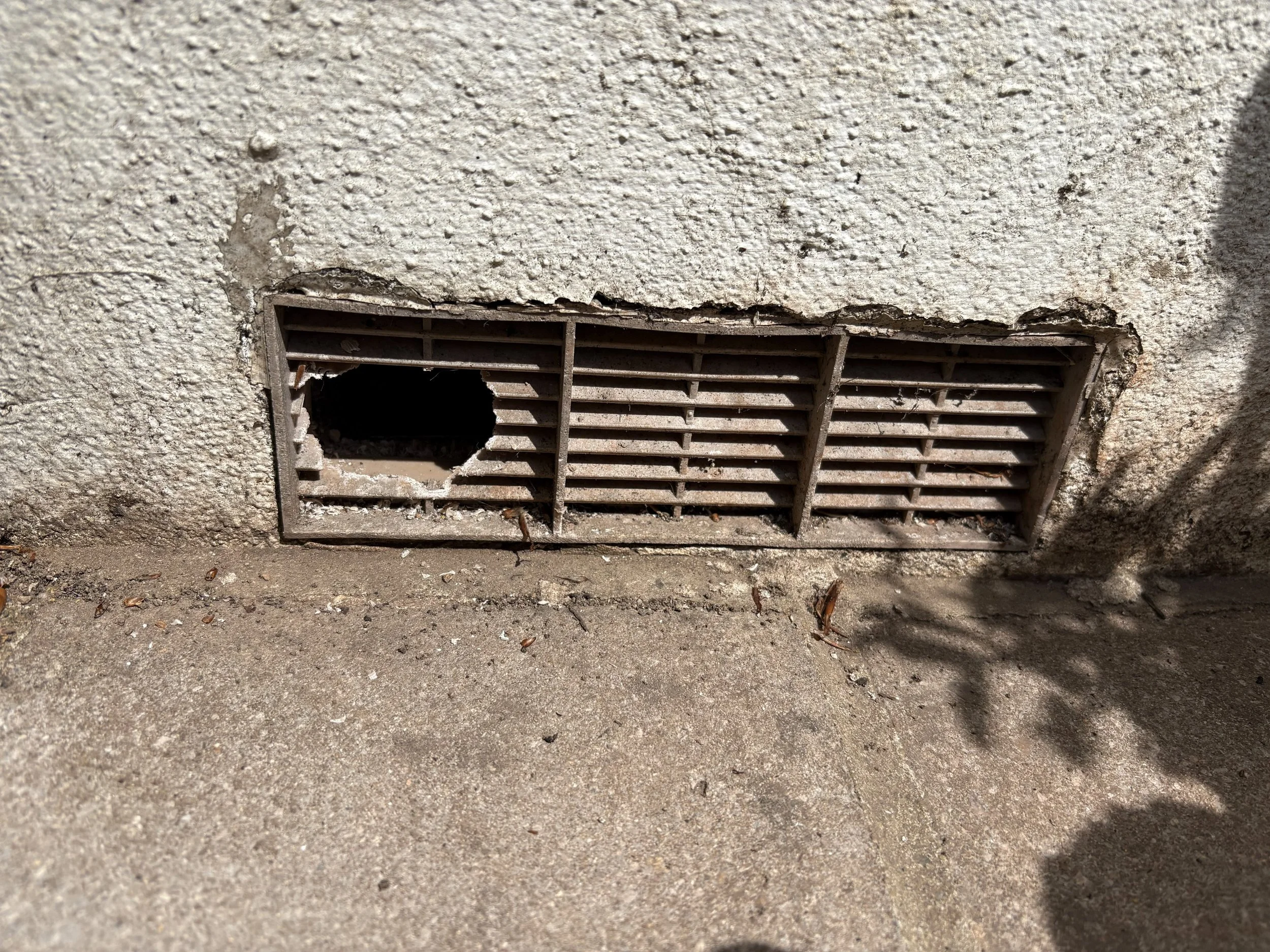 The image shows a damaged ventilation grate on a concrete wall, with one of the horizontal slats broken and a hole in the grate.