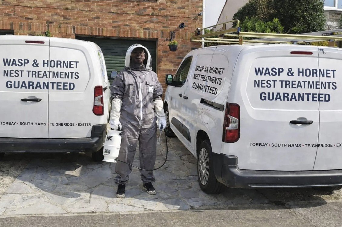 A pest control worker in protective gear standing between two white vans with signs advertising wasp and hornet nest treatments.