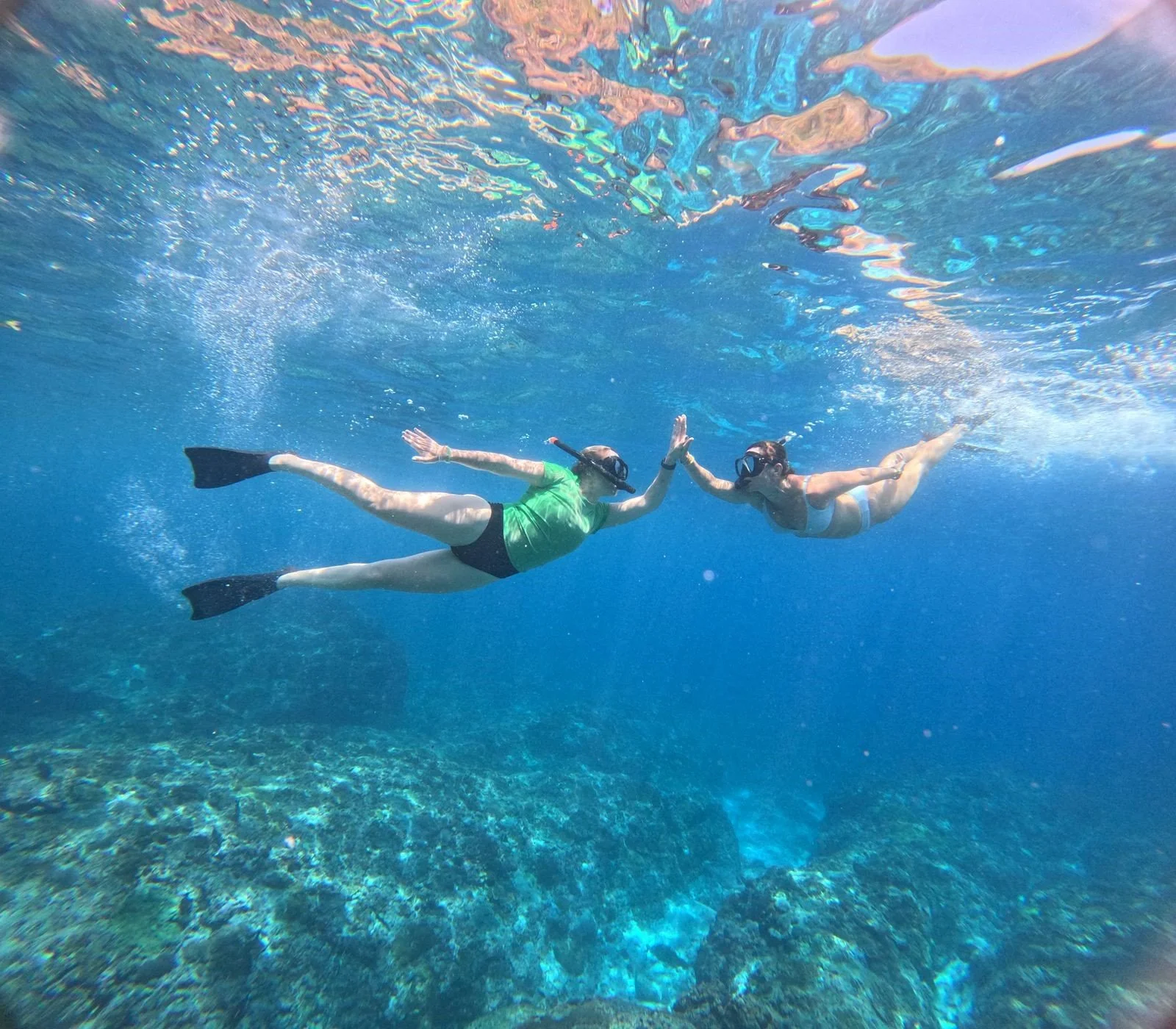 Two women snorkeling underwater, giving each other a high five with a coral reef visible below.
