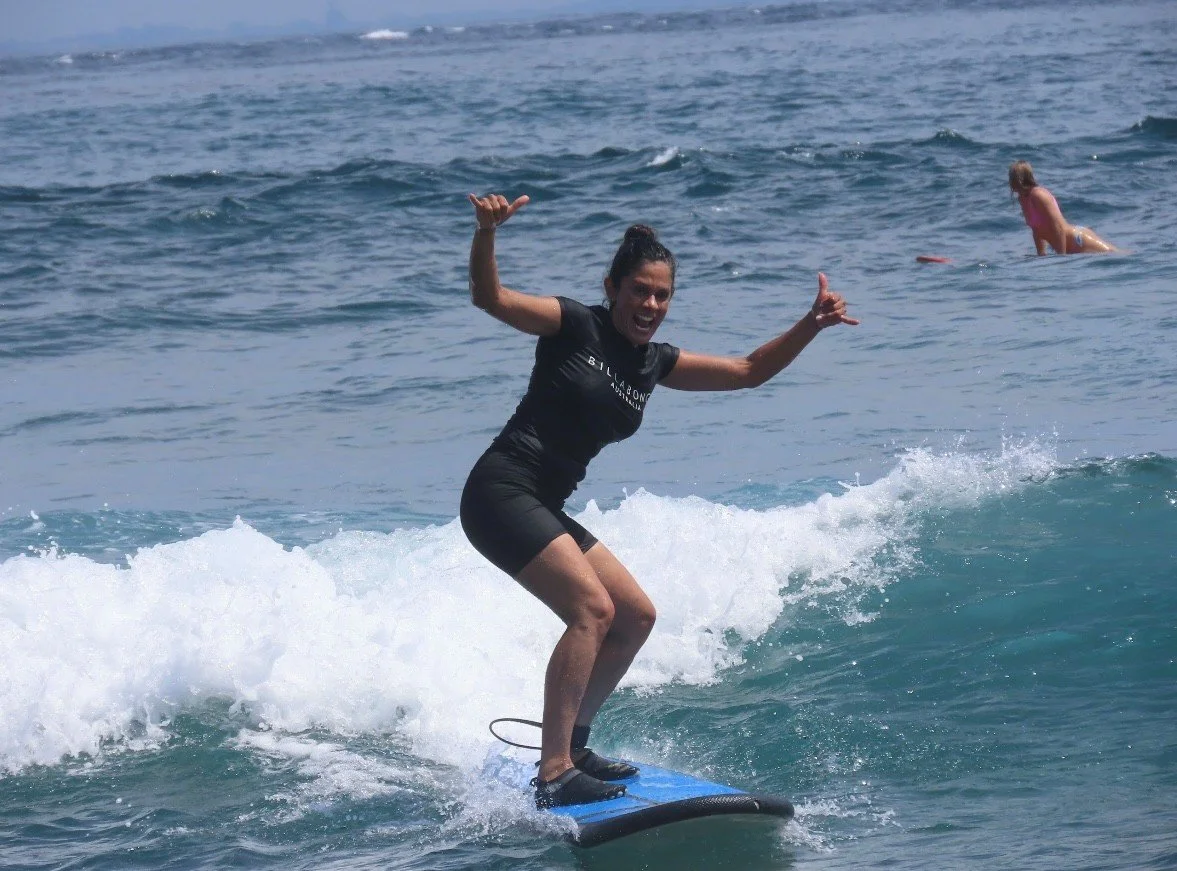 A woman surfing on a wave in the ocean, smiling with her arms raised.