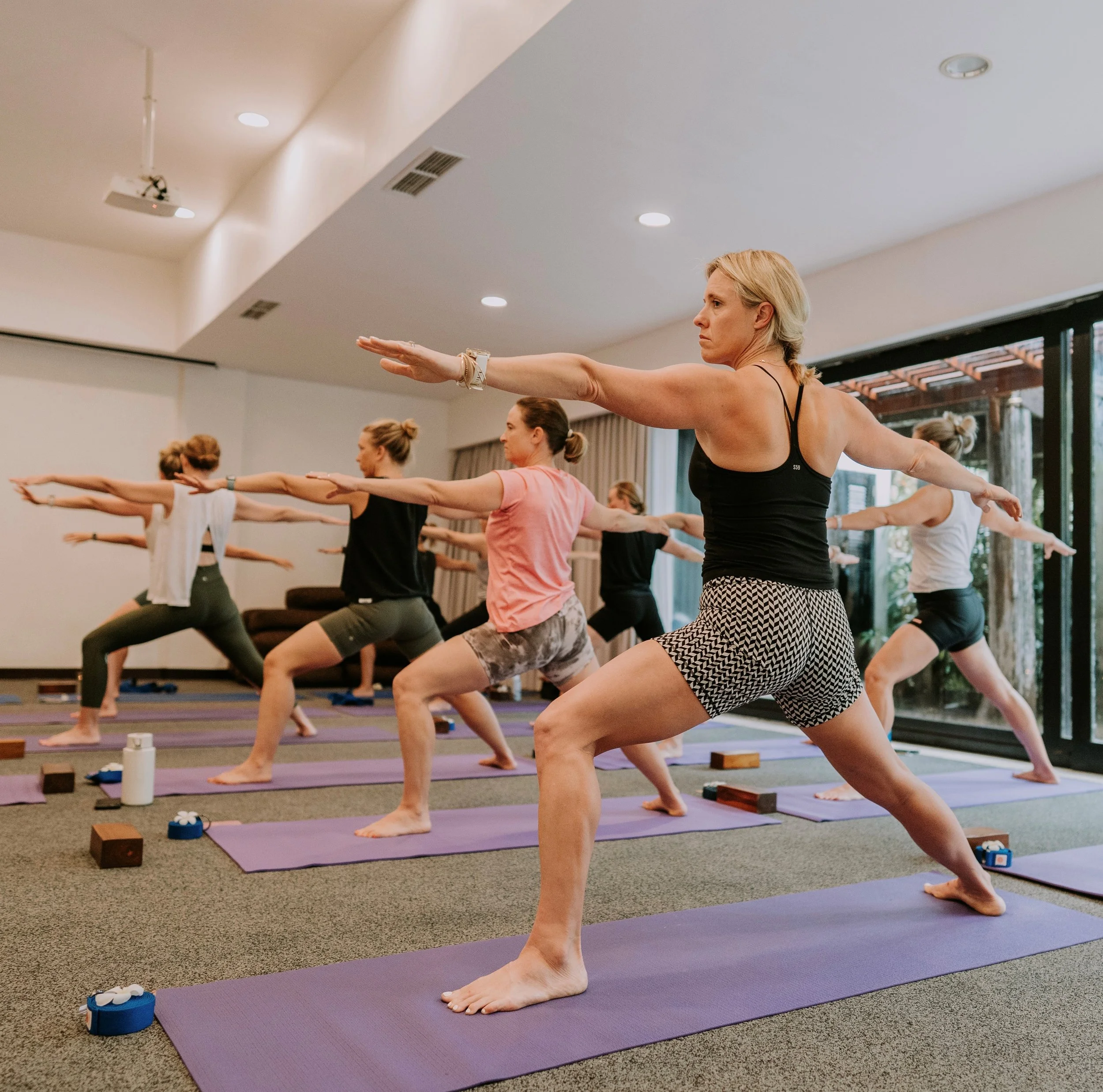 Group of women practicing yoga in a bright indoor studio with large windows, all in a warrior pose on purple mats.