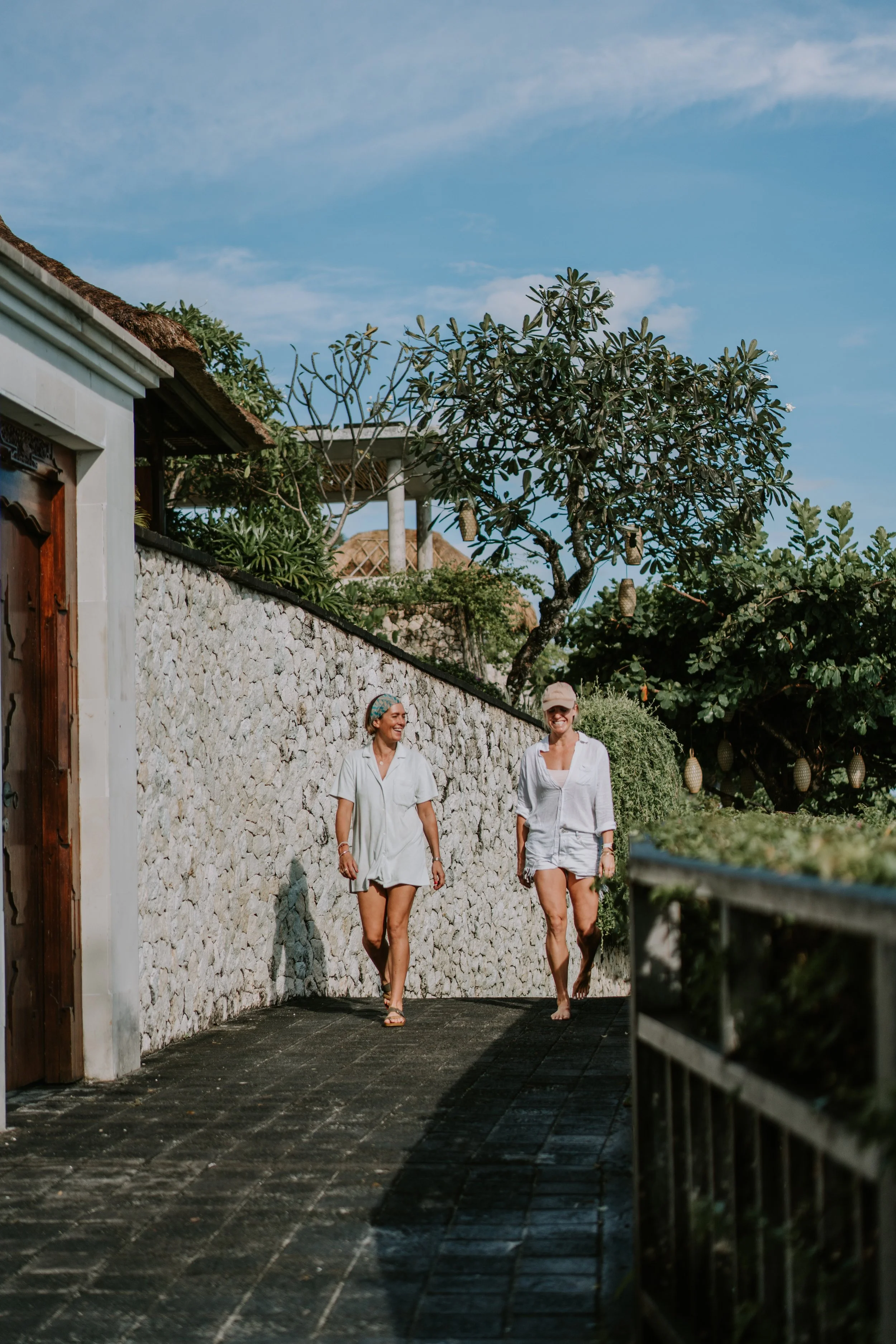 Two women wearing white robes walking barefoot on a stone pathway outdoors under a blue sky with trees and a stone wall in the background.