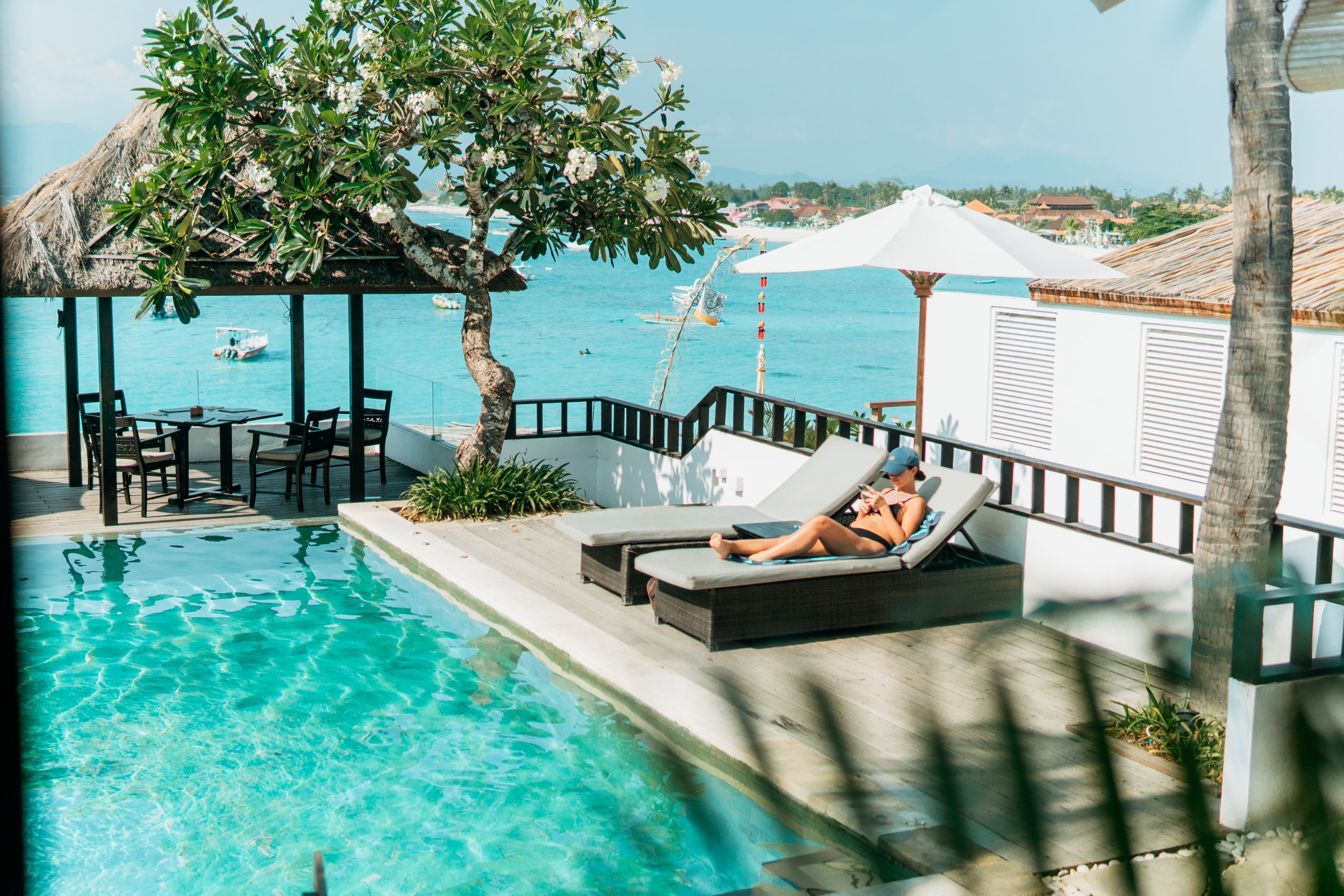 A woman in a swimsuit and cap is relaxing on a lounge chair by a swimming pool, with the ocean in the background. There are umbrellas, trees, and boats visible in a sunny, tropical setting.