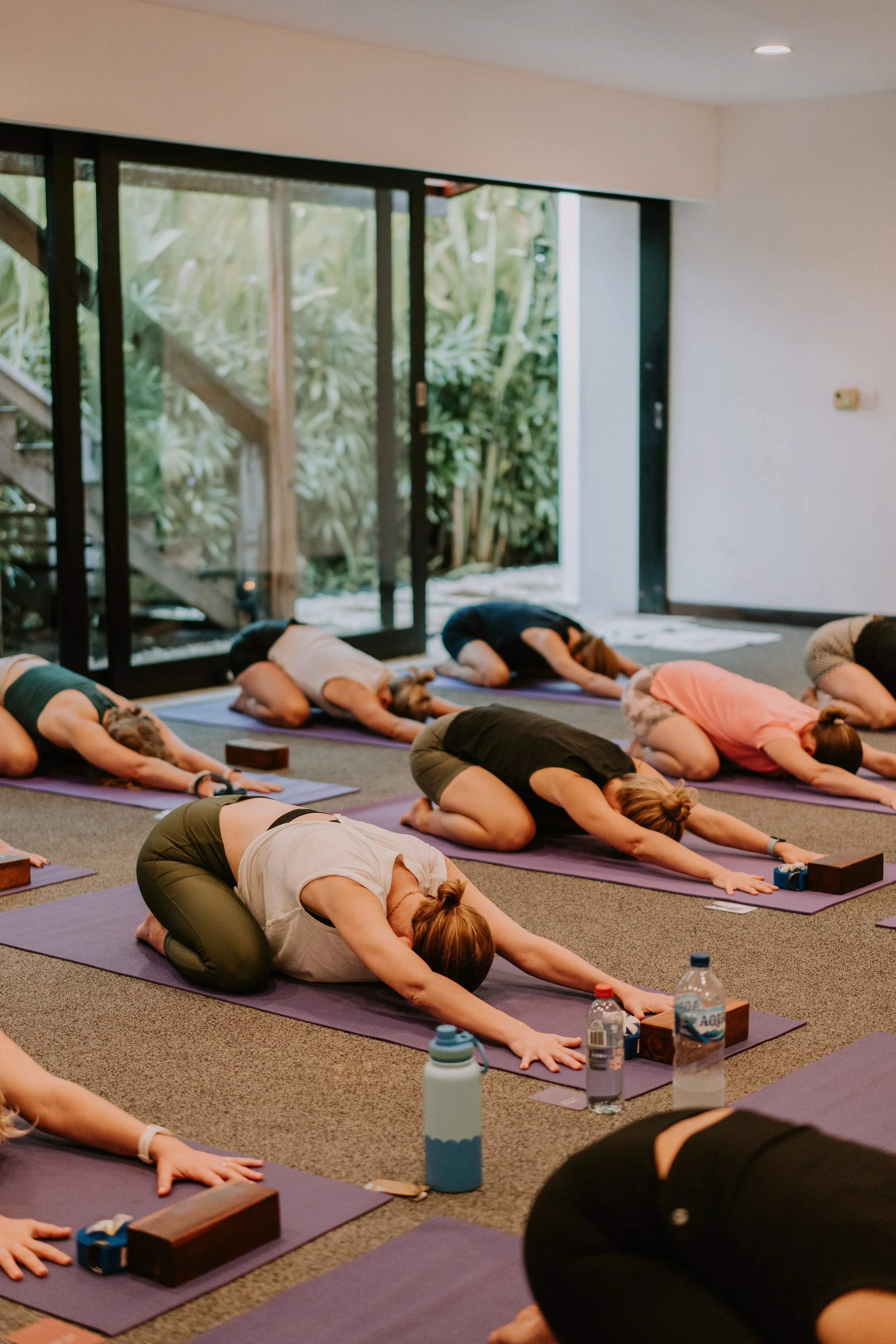 People participating in a yoga class, performing child's pose in a spacious indoor studio with large glass doors opening to greenery outside.