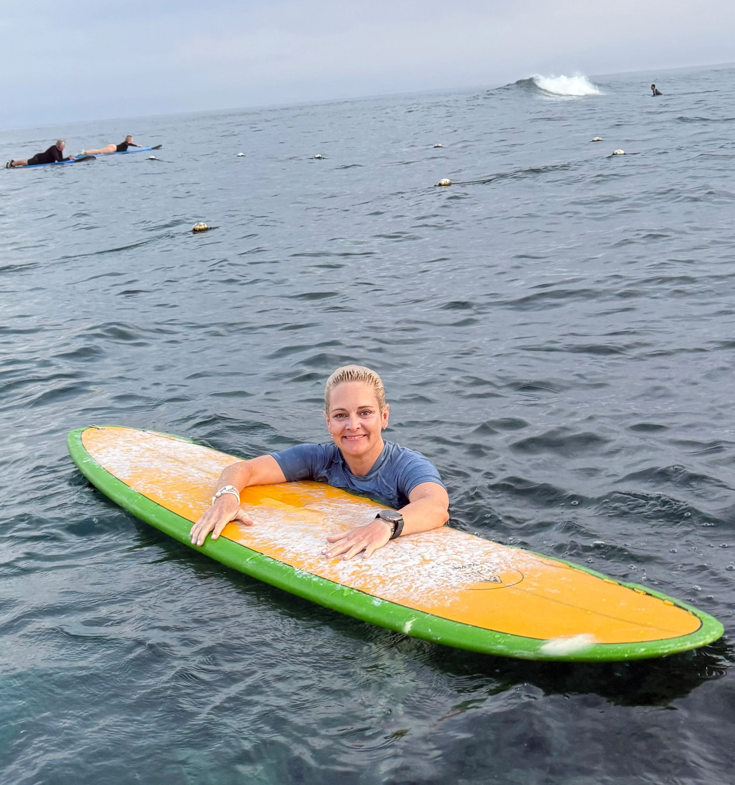 Woman smiling on a surfboard in the water at the beach with other surfers in the background.