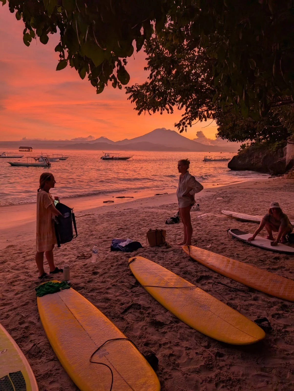 People preparing for paddleboarding on a beach during sunset with a volcano in the background.