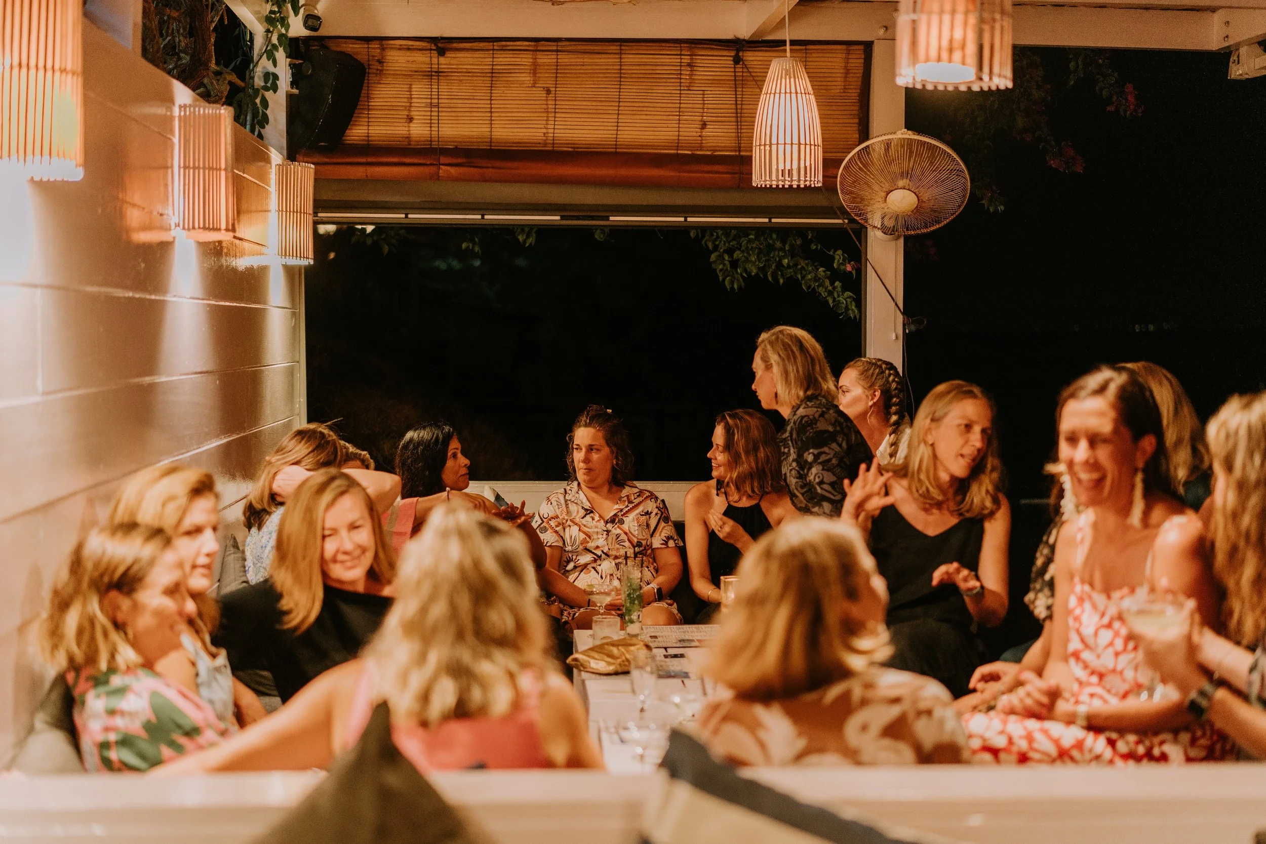 Group of women socializing at a dinner party in a warmly lit outdoor setting at night.