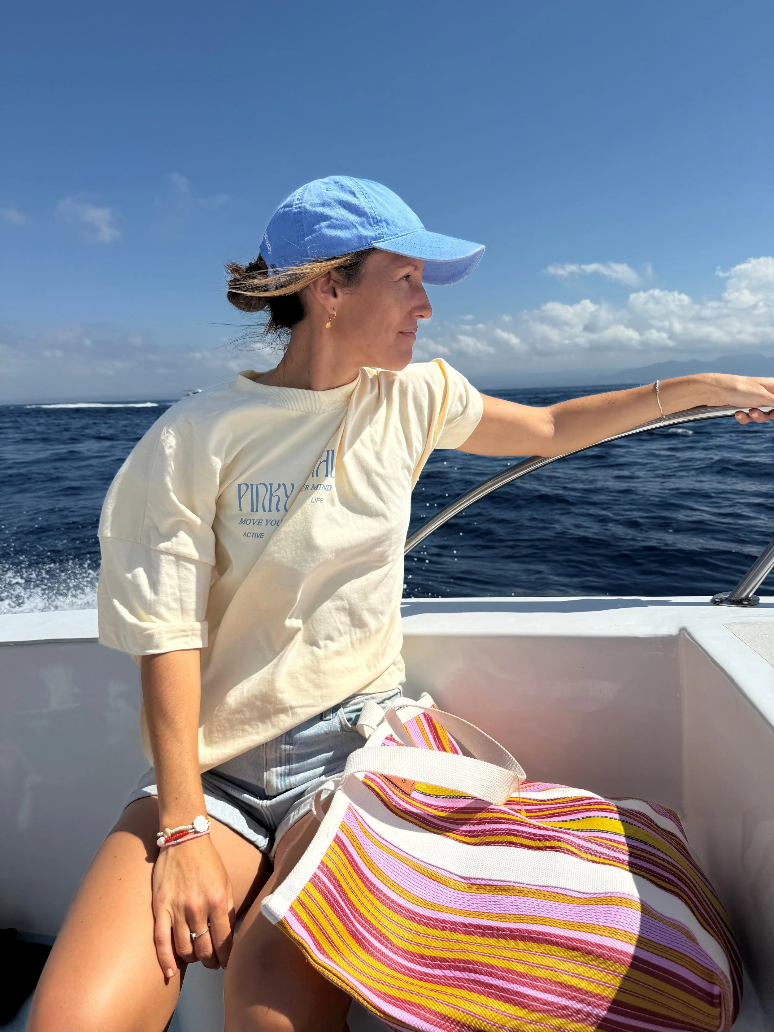A woman with light brown hair in a bun, wearing a blue cap, a cream-colored T-shirt, and denim shorts, sitting on a boat and looking to the left with an ocean background and blue sky.