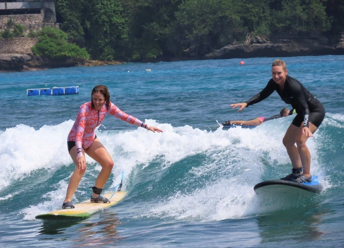 Two women surfing on small waves in a body of water, with one woman in a pink and orange patterned long sleeve shirt and the other in a black wetsuit, smiling and balancing on surfboards, with a backdrop of trees and rocky shoreline.