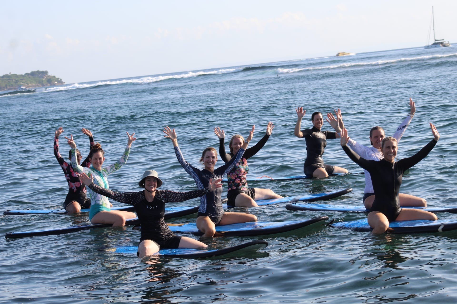 Group of seven women practicing stand-up paddleboarding in the ocean, raising their arms and smiling, with a distant landmass and sailboats in the background.