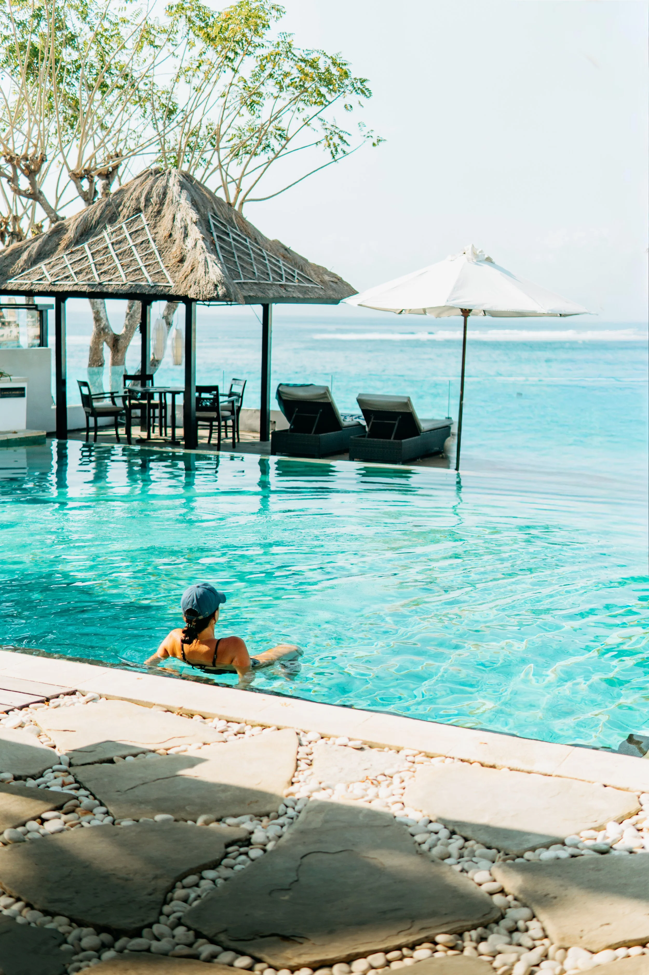 A person sitting in a swimming pool by the beach with a thatched-roof hut, lounge chairs, a white umbrella, and ocean view in the background.