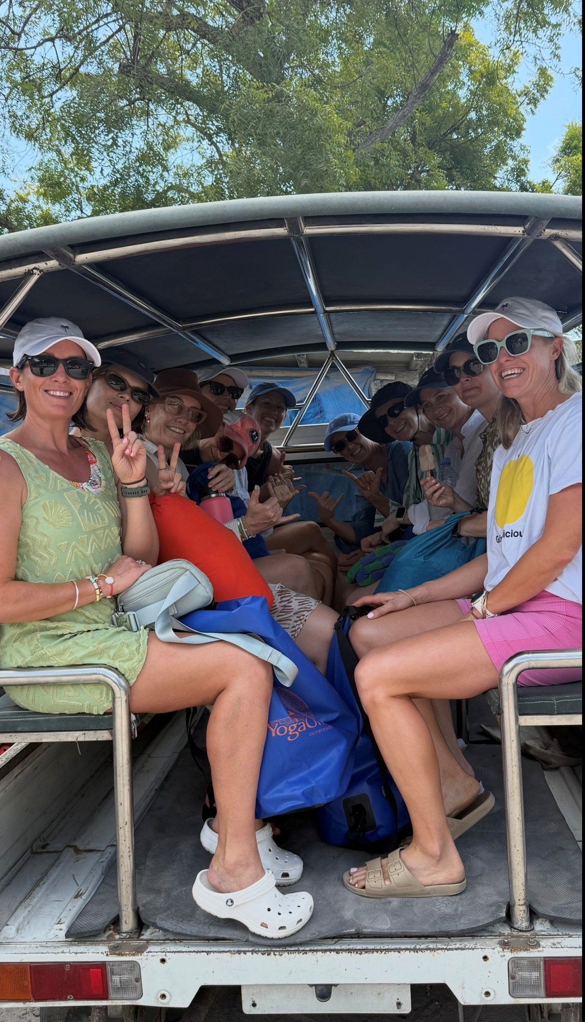 Group of women sitting in a boat, smiling, wearing sunglasses and hats, with a clear sky and green trees in the background.