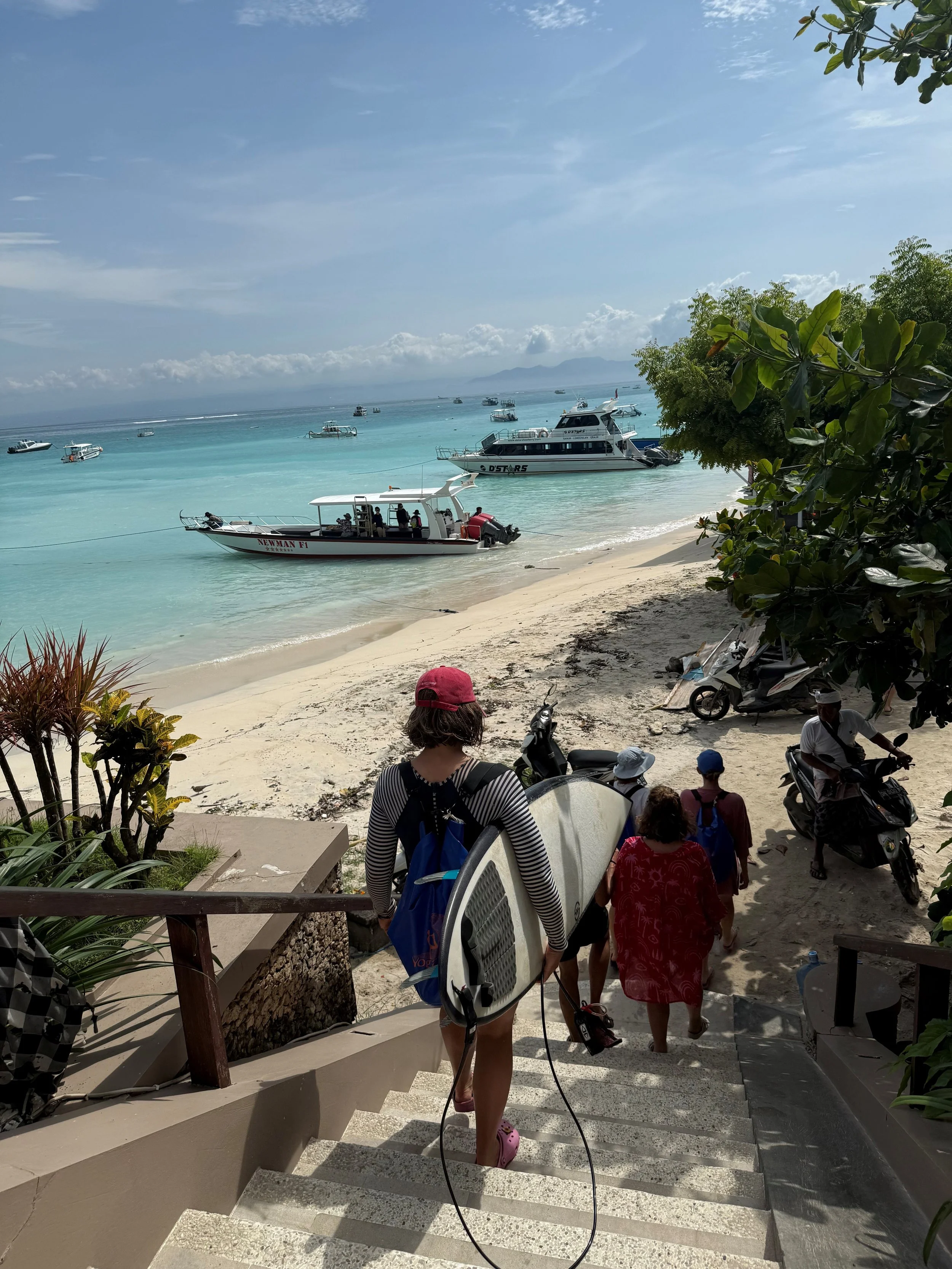Tourists descending steps to a beach with boats anchored in turquoise water, palm trees, and a cloudy sky.