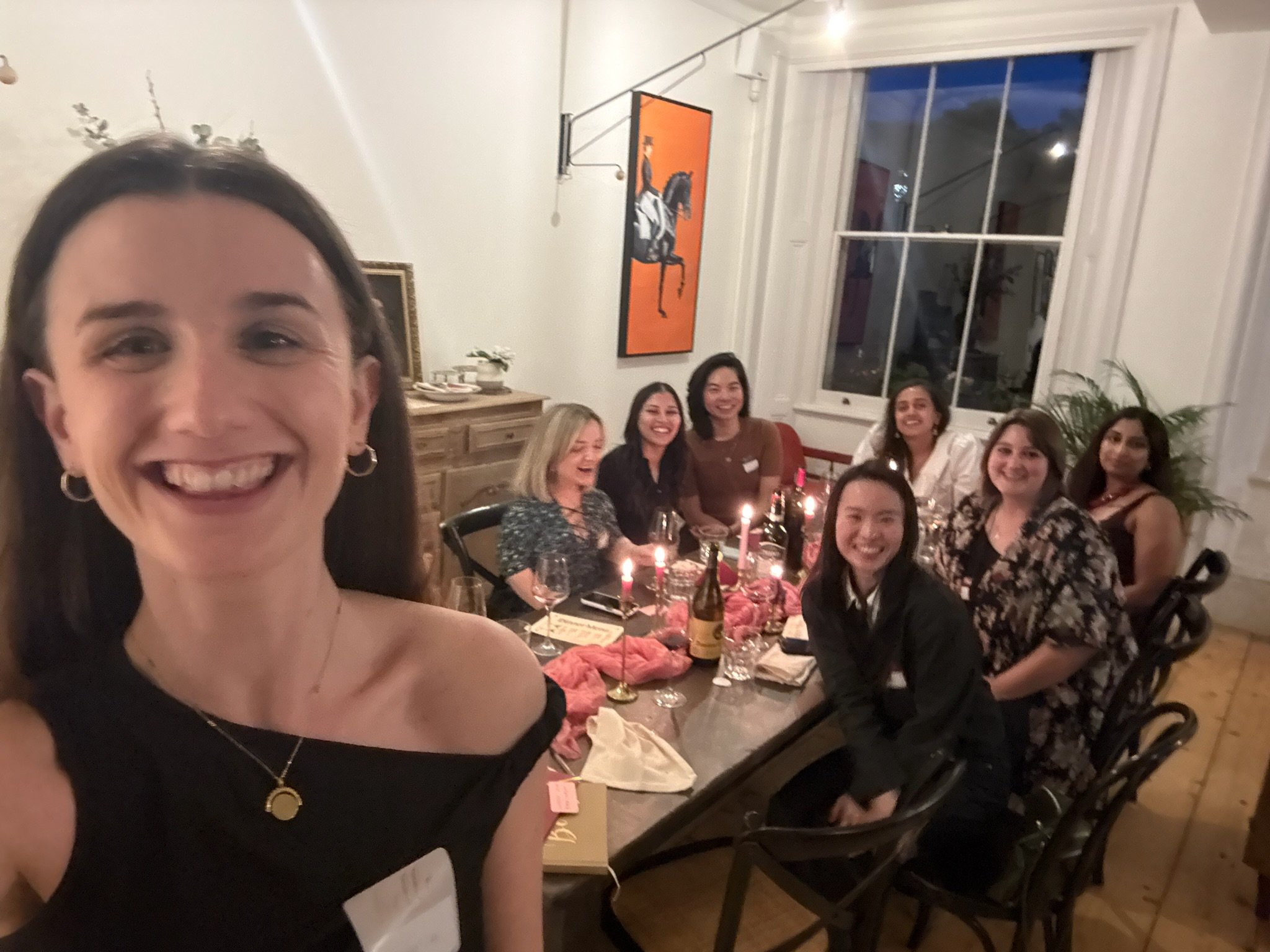 Group of women at a dinner party smiling and celebrating, with candles, food, and drinks on the table, in a well-lit room with artwork and a window.