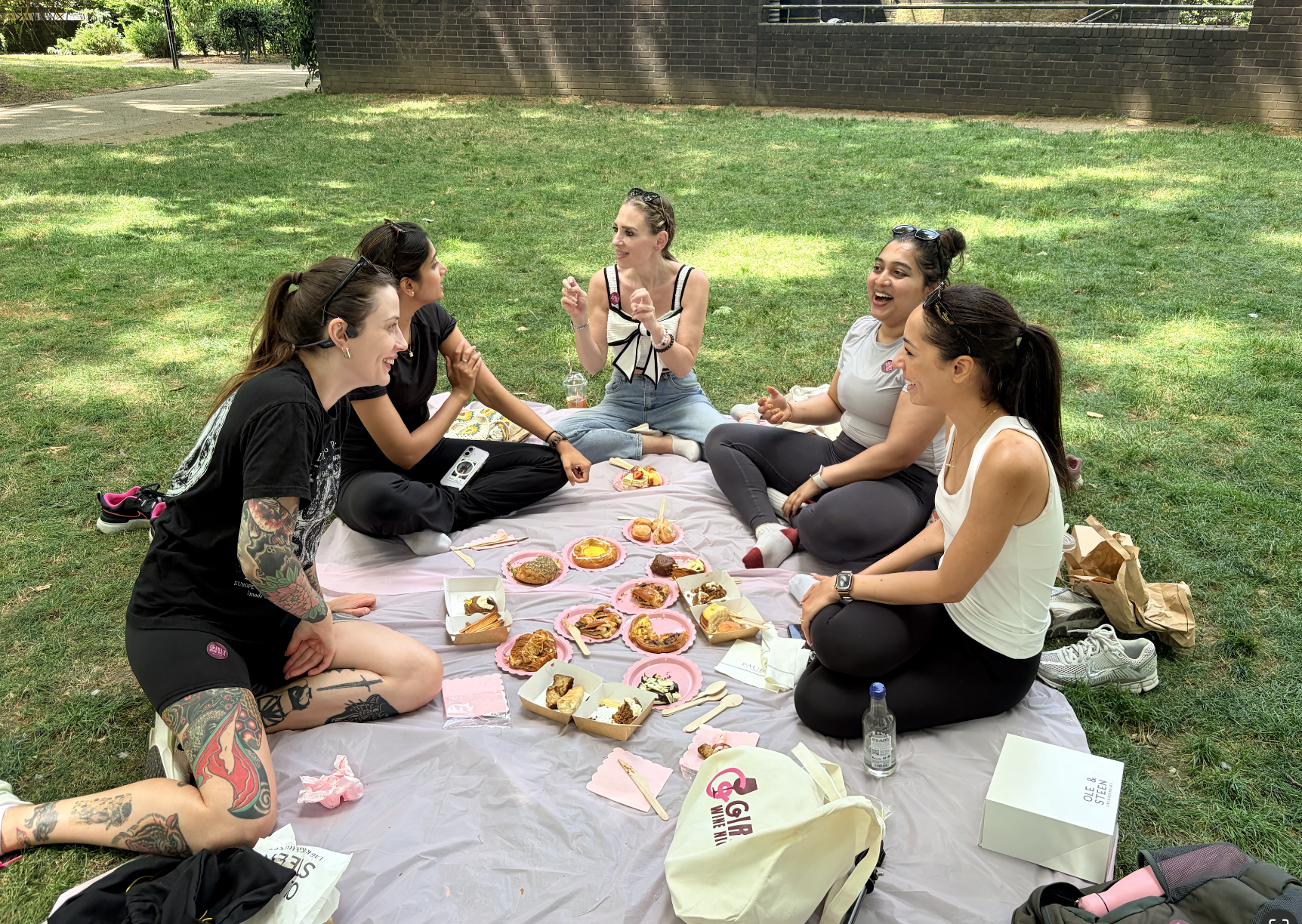 A group of six women sitting on a picnic blanket outdoors, having a picnic with various cakes, pastries, and snacks. They are smiling and engaging in conversation on a sunny day with trees and a brick wall in the background.