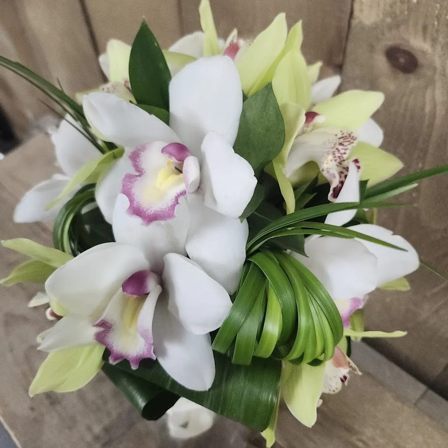 Bouquet of white orchids with pink and yellow centers, accented with green leaves and grass-like foliage, set against a wooden background.