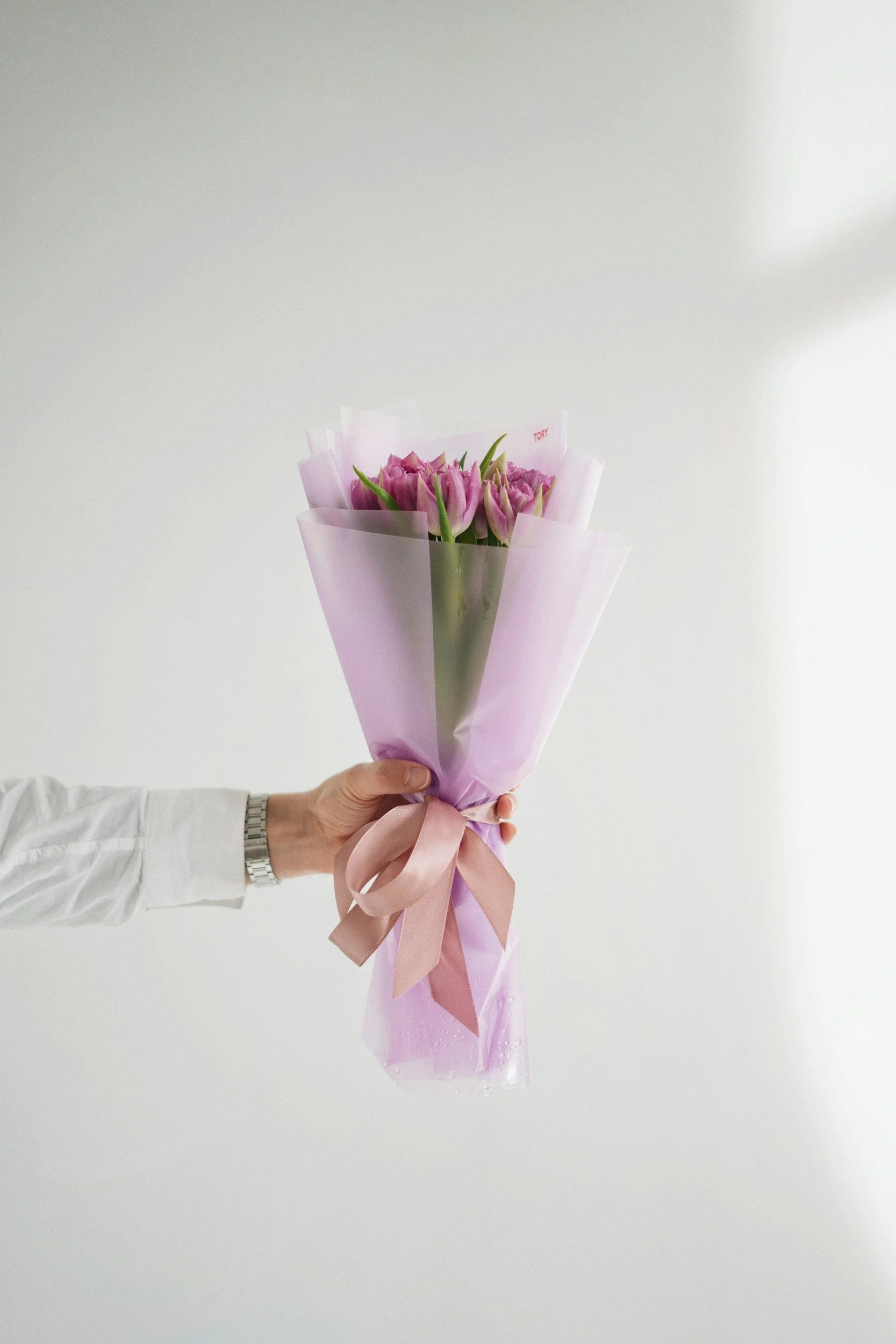 Person holding a bouquet of pink tulips wrapped in pink and purple cellophane with a pink ribbon.