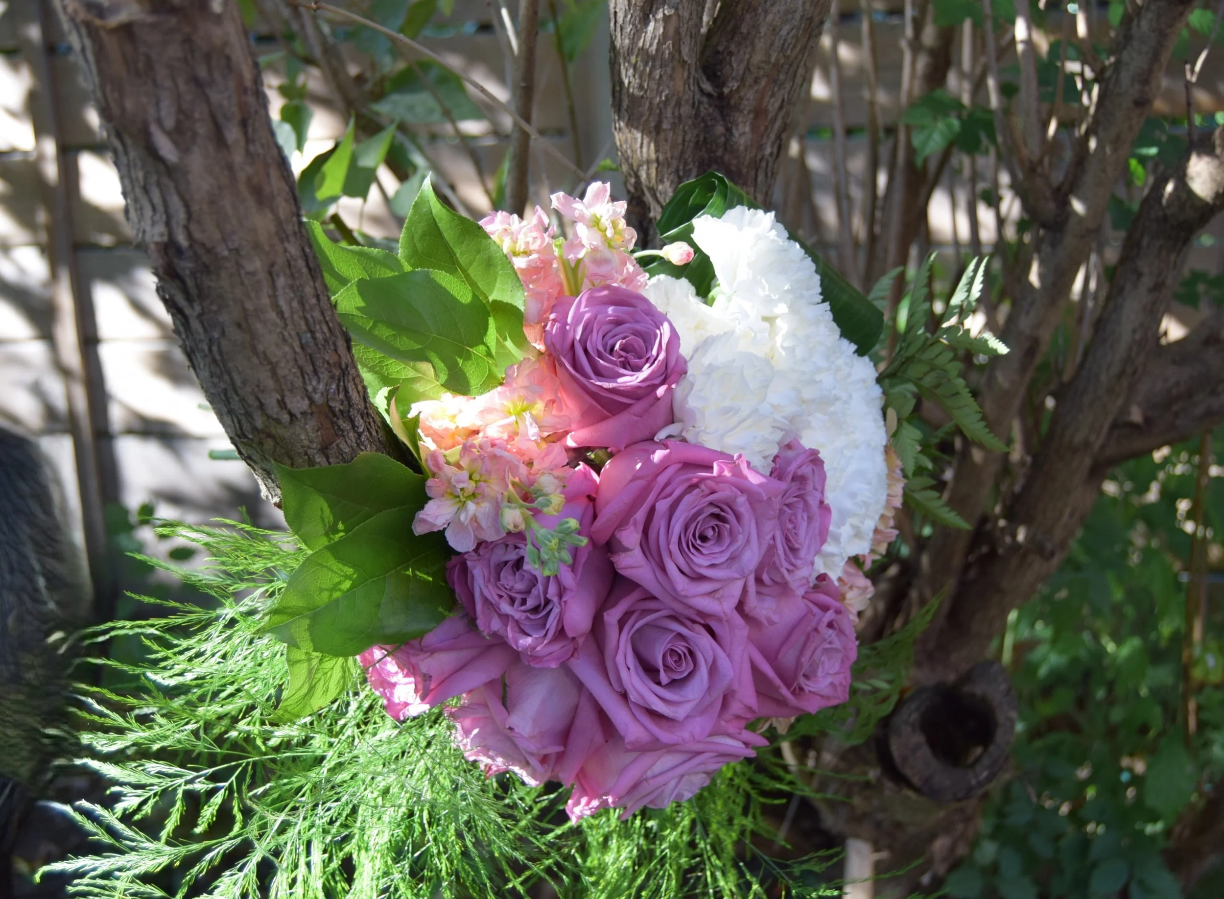 A bouquet of pink, purple, and white flowers including roses and hydrangeas, attached to a tree with green leaves and a background of branches and foliage.