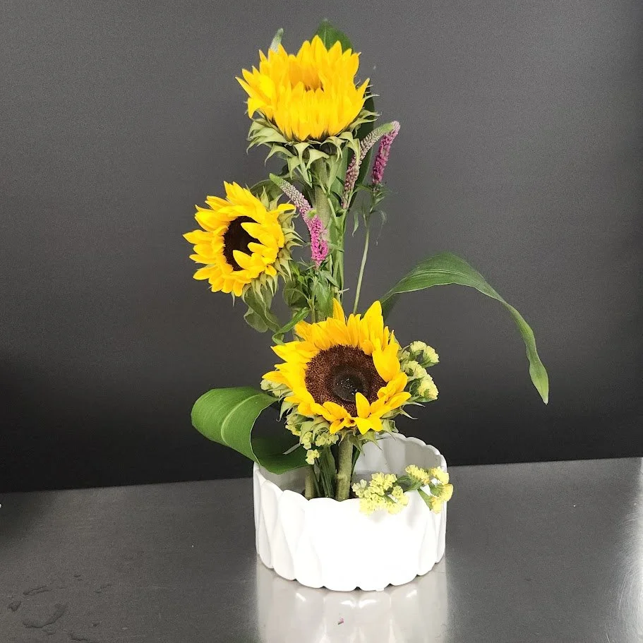 A floral arrangement of sunflowers and other flowers in a white vase on a dark surface with a gray background.