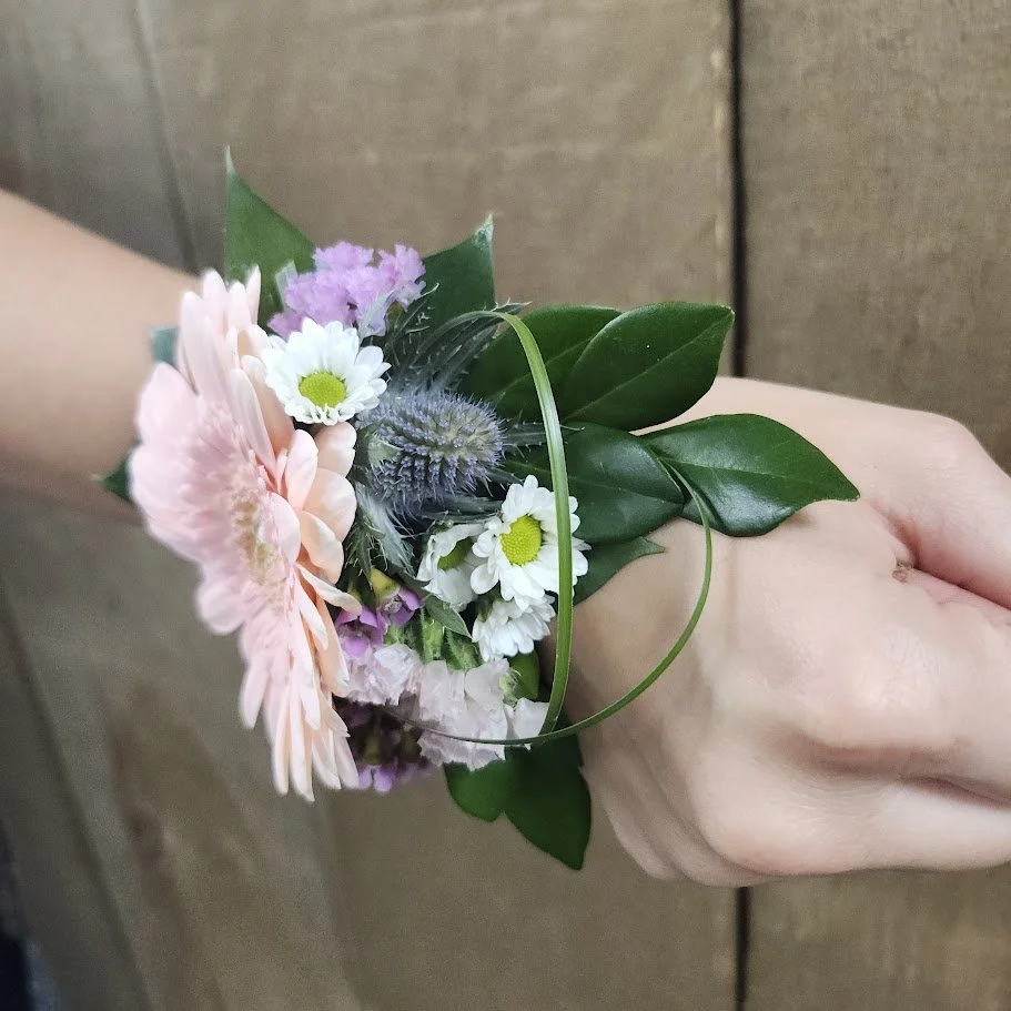 A person's hand holding a small bouquet of flowers with pink, white, and purple blooms along with green leaves.