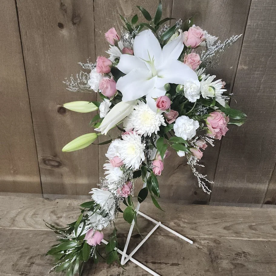 A floral arrangement on a white stand includes white lilies and orchids, pink roses, and various greenery against a wooden background.
