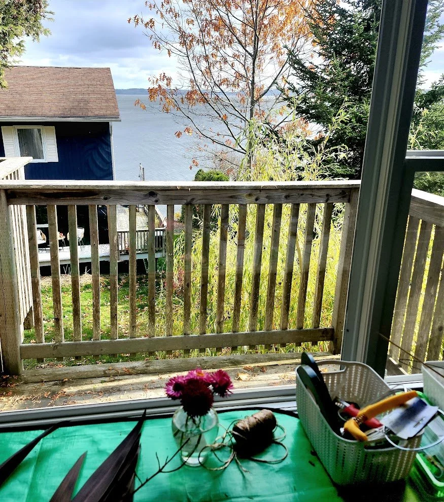 View from inside a house showing a small balcony with a wooden railing, overlooking a lake and trees with fall foliage, and part of a neighboring house with a brown roof.