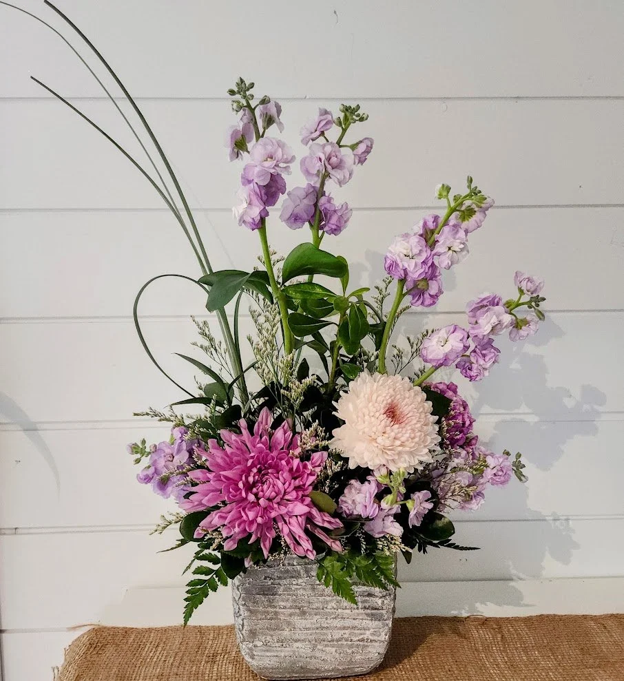 Flowers in a white vase with pink roses, pink asters, white baby's breath, and greenery, placed against a white wooden wall.