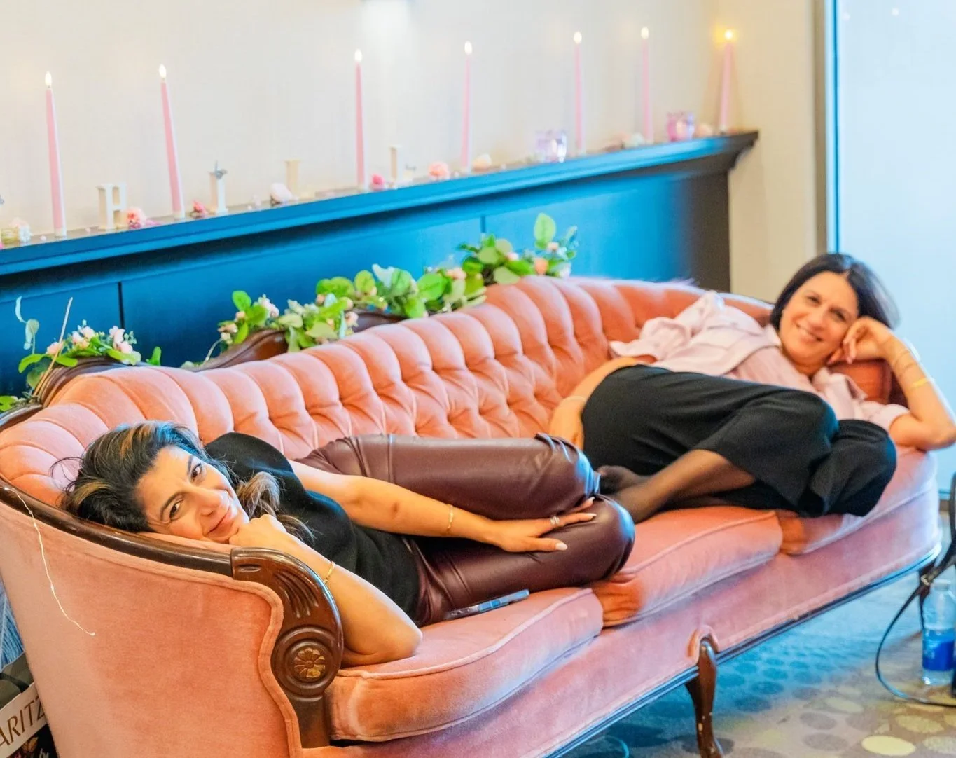 Two women lie on a pink vintage-style couch with a guernsey back, green floral decorations on top, and pink candles behind them in an indoor setting, smiling at the camera.