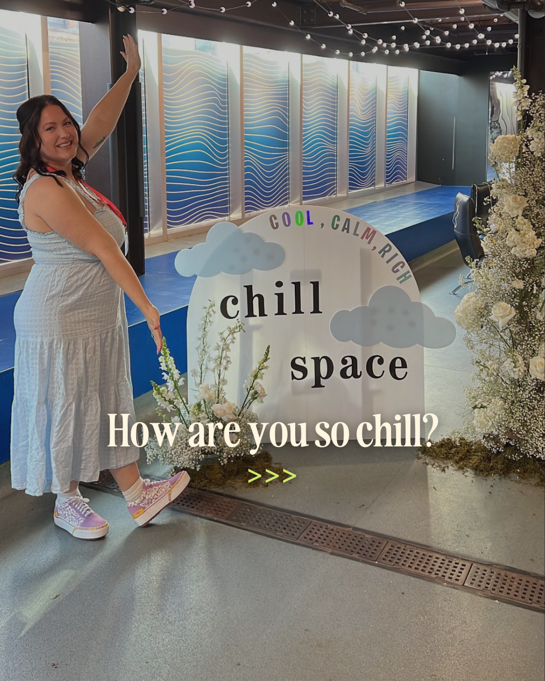 A woman in a white dress and colorful sneakers smiling and pointing at a sign that reads 'chill space' with clouds and the words 'cool, calm, rich' above it, surrounded by floral arrangements and a decorated stage in the background.