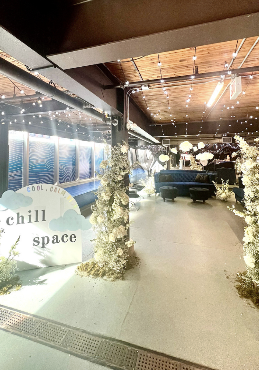 Interior of a lounge or chill space decorated with string lights, floral arrangements, and cloud-shaped decor. There's a sign that says 'chill space' and a small stage or platform in the background.