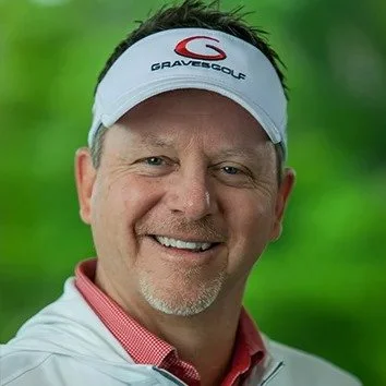 Close-up of a man smiling outdoors, wearing a white golf visor and a white shirt with a red collar, with green foliage in the background.