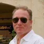 A man wearing sunglasses and a white shirt standing outdoors.