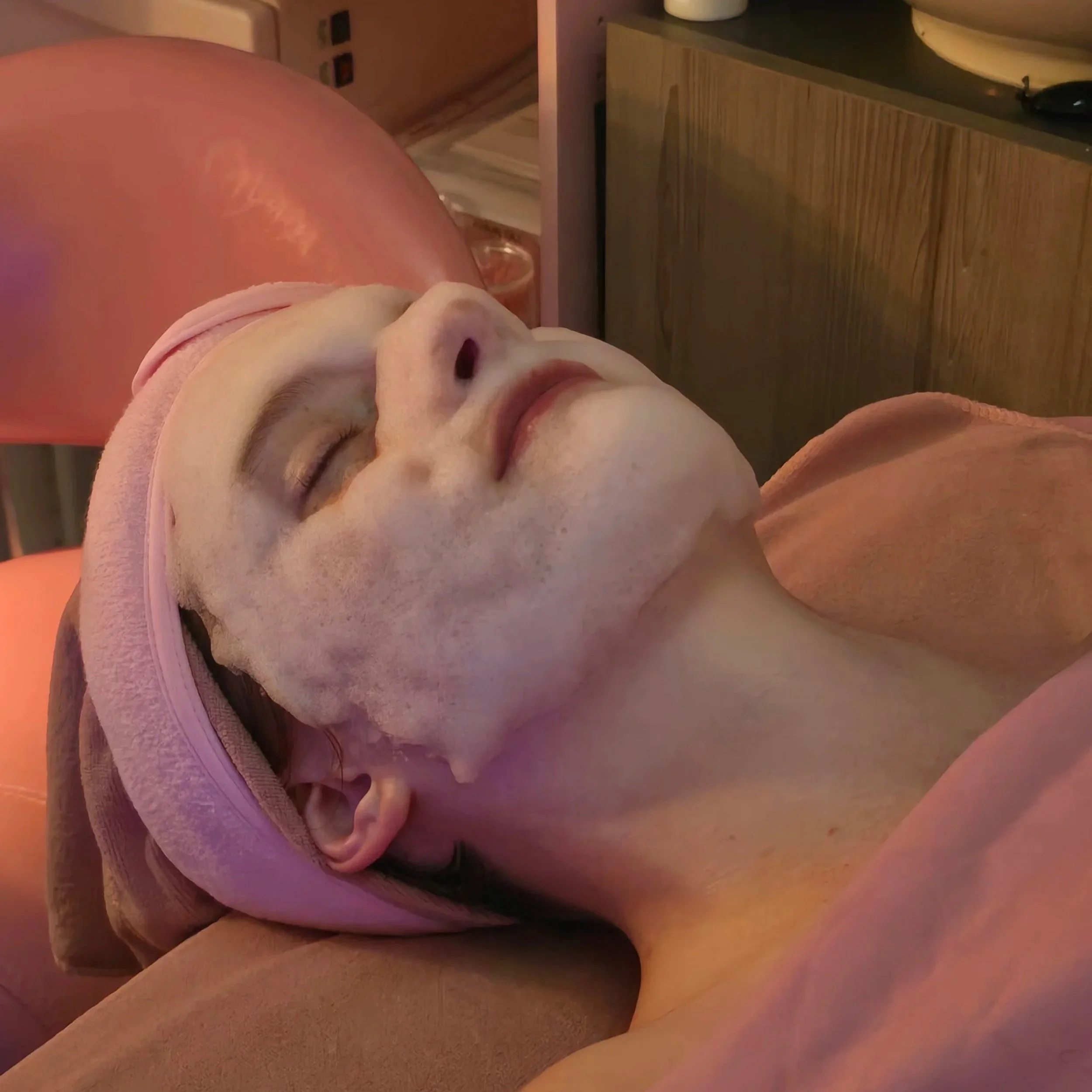 A woman lying down with her eyes closed, wearing a pink headband and facial mask, in a relaxing skincare setting.
