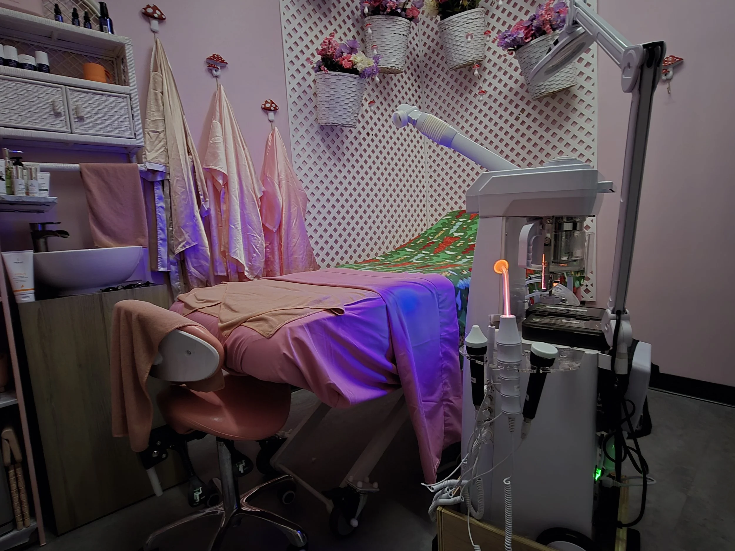 Facial treatment room with a bed covered in pink and green sheets, a multi-functional facial technique machine, hanging towels, shelves with books and supplies, and decorative floral baskets on the lattice background wall.